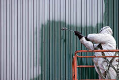 Pintor profesional en Drummondville, con equipo de protección completo, sobre un cubo. Pinta el revestimiento de un edificio metálico.