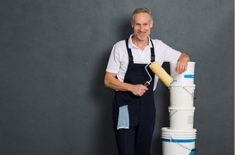 Mature smiling professional painter in Drummondville with a roller in his right hand and his left arm resting on paint boilers. 